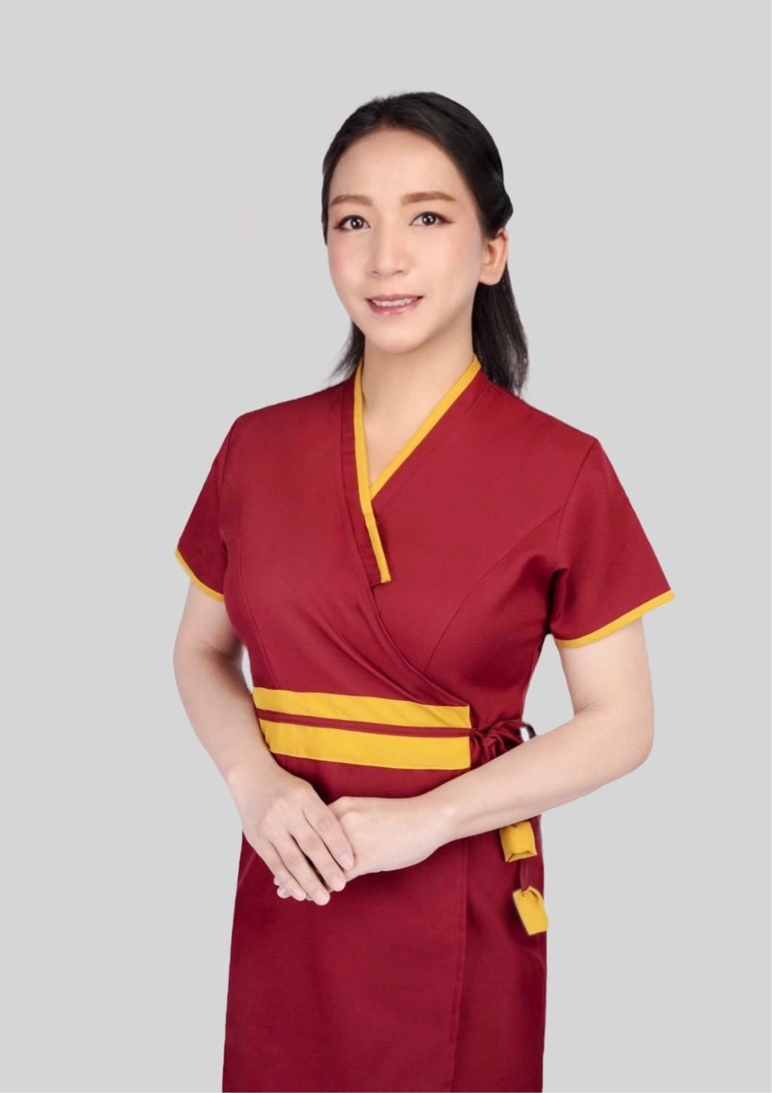 A transgender massage therapist with long, black hair, half-tied back, wearing a red massage uniform, stood with her hands clasped in front of her, smiling.
