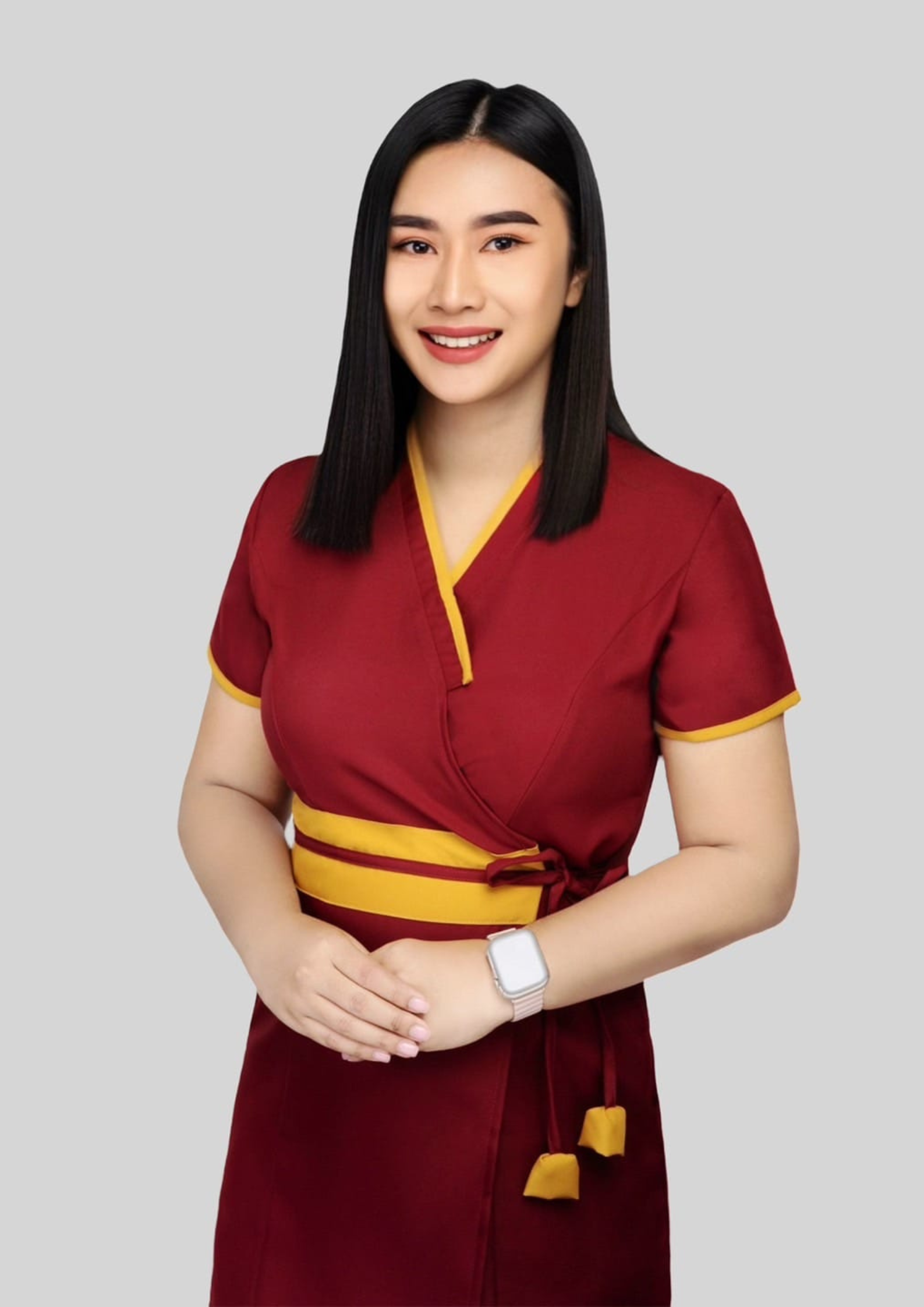 A ladyboy massage therapist smiled, wearing a red massage uniform and a pink watch, and stood with her hands clasped in front of her. by The Elite Bangkok Thai Massage